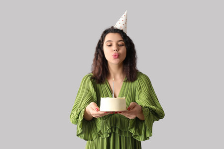 Beautiful young woman in party hat with sweet cake celebrating Birthday on gray background