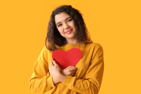 Beautiful young woman with red paper heart on yellow backgroundの写真素材