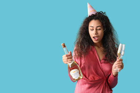 Beautiful young African-American woman in party hat with glass and bottle of champagne celebrating Birthday on blue backgroundの写真素材