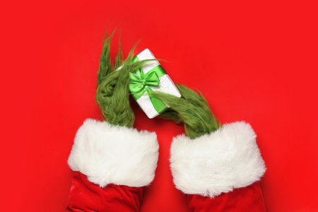 Green hairy hands of creature in Santa costume with gift box on red backgroundの写真素材