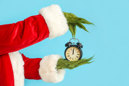 Green hairy hands of creature in Santa costume with alarm clock on blue background