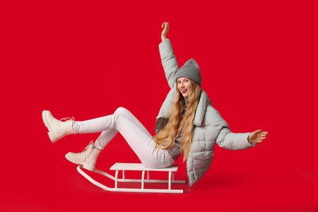 Beautiful young happy woman in warm winter clothes with sledge on red background