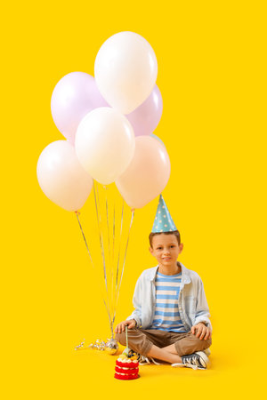 Cute little boy with Birthday cake and balloons sitting on yellow background