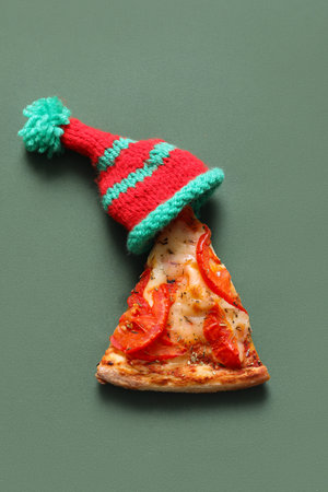Piece of delicious pizza and Christmas knitted hat on green background