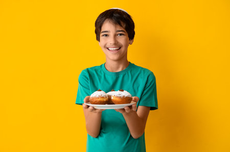 Little boy in kipa holding plate with tasty donuts on yellow background. Hanukkah celebrationの写真素材