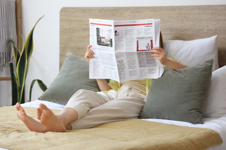 Beautiful young woman reading newspaper in bedroom at homeの写真素材