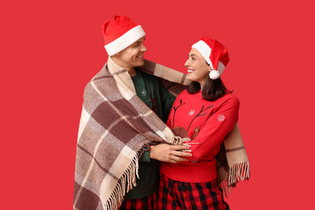 Happy young couple in Christmas pajamas and Santa hats wrapped in plaid on red backgroundの写真素材