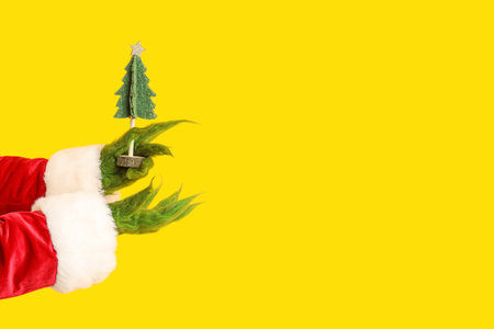 Green hairy hands of creature in Santa costume with decorative fir tree on yellow backgroundの写真素材