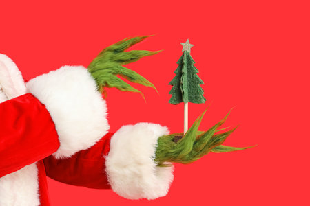 Green hairy hands of creature in Santa costume with decorative fir tree on red backgroundの写真素材