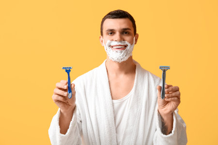 Handsome young man with razors and shaving foam on his face against yellow backgroundの写真素材