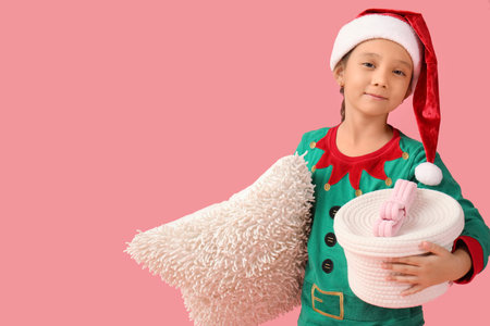 Cute little girl in elf costume with basket and cushion on pink background