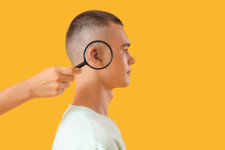 Woman magnifying teenage boy's ear with hearing aid on yellow backgroundの写真素材