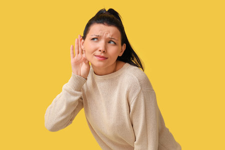 Young woman trying to hear something on yellow backgroundの写真素材