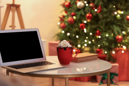 Cup of hot chocolate with marshmallows near laptop on coffee table in festive living roomの写真素材