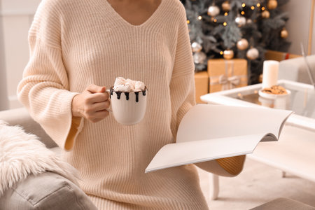 Woman holding cup of hot chocolate with marshmallows while reading magazine in festive living roomの写真素材