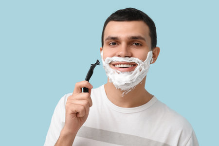 Handsome young man with razor and shaving foam on his face against blue backgroundの写真素材
