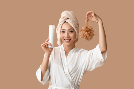 Young Asian woman in bathrobe with shower gel and loofah on brown backgroundの写真素材