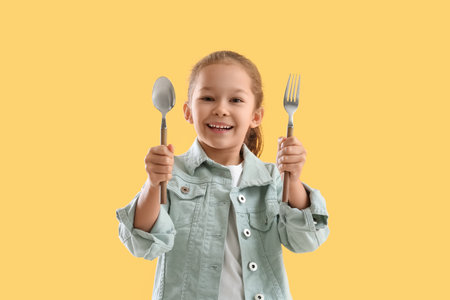 Cute little girl with fork and spoon on yellow backgroundの写真素材