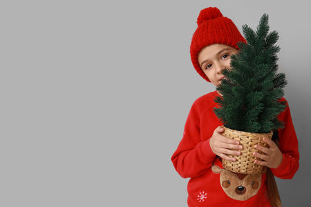 Cute little girl wearing warm hat with Christmas tree in pot on gray backgroundの写真素材
