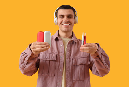 Young man with electronic cigarettes and headphones on yellow background