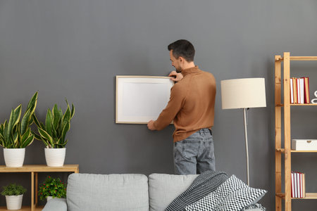 Handsome man hanging photo frame on dark wall at home, back viewの写真素材