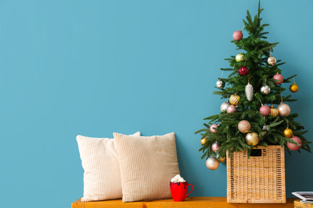 Beautiful Christmas tree, cup of cocoa drink and cushions on wooden bench against color wallの写真素材