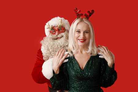 Santa Claus and beautiful mature woman in Christmas deer horns on red backgroundの写真素材
