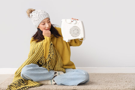 Frozen young Asian woman in winter clothes with electric heater near light wallの写真素材