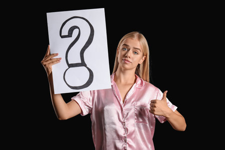 Young woman in pajamas holding paper with question mark and showing thumb-up on black backgroundの写真素材