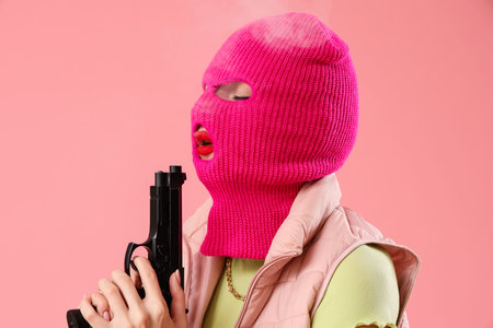 Beautiful young stylish woman in balaclava with gun on pink backgroundの写真素材
