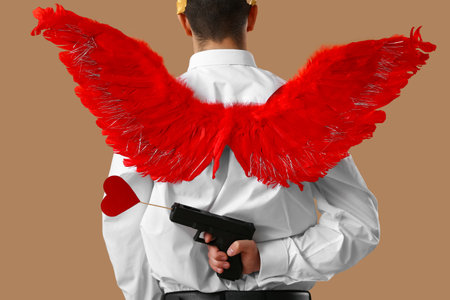 Cool young man dressed as Cupid with gun on beige background, back view. Valentine's Day celebrationの写真素材