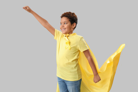 Cute little African-American boy in superhero costume with yellow ribbon on gray background. Childhood cancer awareness conceptの写真素材