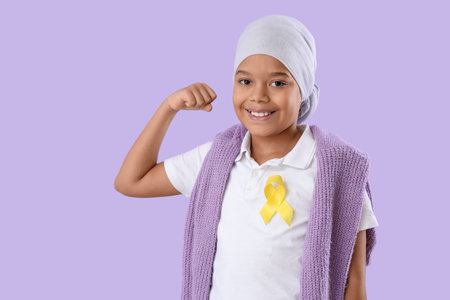 Cute little African-American boy after chemotherapy with yellow ribbon showing muscles on lilac background. Childhood cancer awareness conceptの写真素材