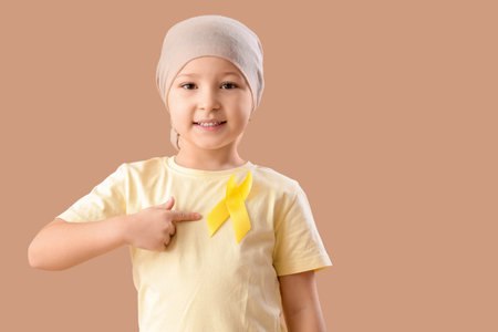 Cute little girl after chemotherapy pointing at yellow ribbon on brown background. Childhood cancer awareness conceptの写真素材