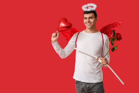 Young man dressed as Cupid with balloon, bow and rose on red background. Valentine's Day celebrationの写真素材