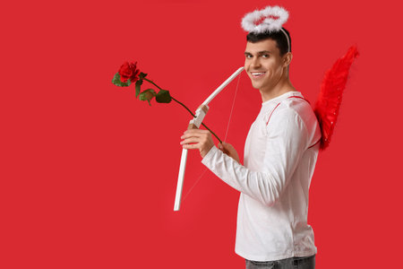 Young man dressed as Cupid with bow and rose on red background. Valentine's Day celebration