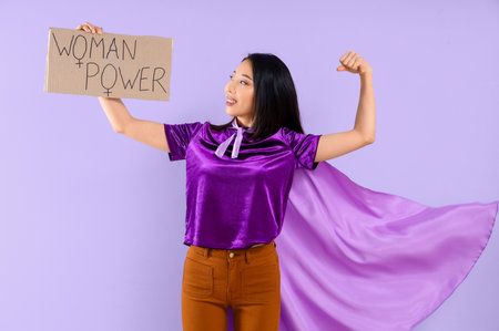 Young Asian girl dressed as superhero holding paper with text WOMAN POWER on lilac backgroundの写真素材