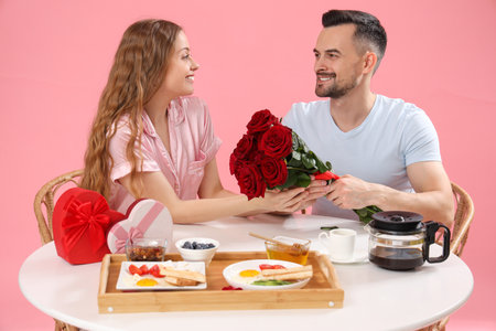 Young couple with roses having breakfast at table on pink background. Valentine's Day celebrationの写真素材