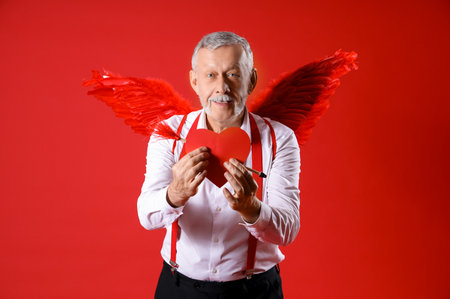 Mature man dressed as Cupid holding heart with arrow on red background. Valentine's Day celebrationの写真素材