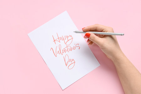 Female hands with red manicure, pen and festive postcard on pink background. Valentine's Day celebrationの写真素材