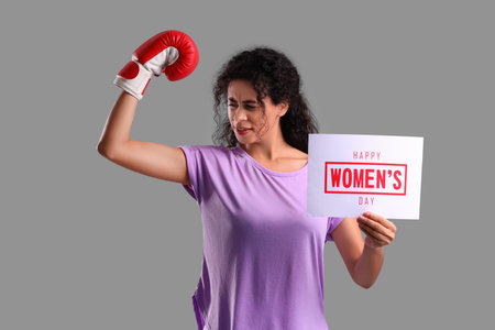 Young African-American woman in boxing glove holding paper sheet with text HAPPY WOMEN'S DAY on gray background. International Women's Day celebrationの写真素材