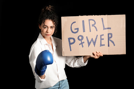 Beautiful young African-American woman in boxing gloves holding paper with text GIRL POWER on black background. Women history monthの写真素材
