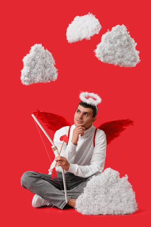 Thoughtful young man dressed as Cupid with bow in clouds on red background. Valentine's Day celebrationの写真素材