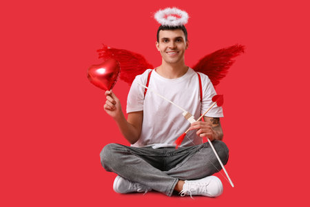 Young man dressed as Cupid with balloon and bow sitting on red background. Valentine's Day celebrationの写真素材