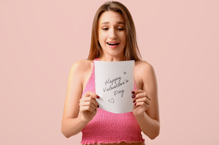 Surprised young woman holding greeting with text HAPPY VALENTINE'S DAY on pink backgroundの写真素材
