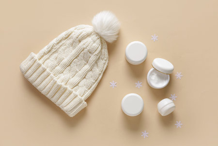Jars of cream with decorative snowflakes and warm winter hat on beige background