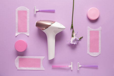 Modern photoepilator with wax strips, razors and jars of cream on purple background