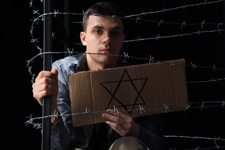Young Jewish man holding paper with David star behind barbed wire on black background. International...