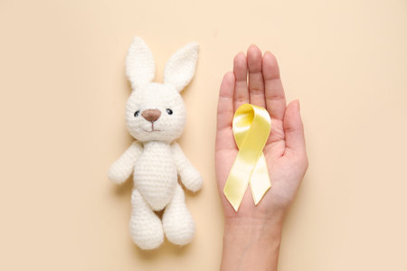Female hand with toy bunny and golden awareness ribbon on beige background. International Childhood Cancer Dayの写真素材