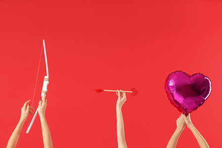 Female hands holding Cupid's bow, arrow and heart-shaped balloon on red background. Valentine's Day celebrationの写真素材